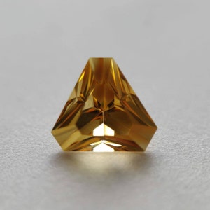May include: A triangular, faceted gemstone with a golden yellow color. The gemstone is cut into a unique shape with many facets that reflect light.