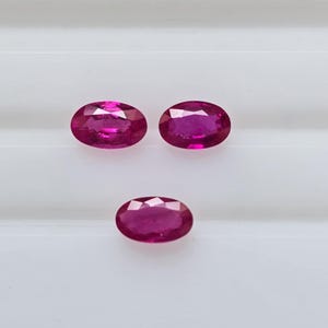 May include: Three oval-cut, vibrant red gemstones. The gems are displayed against a white background, showcasing their polished surfaces and internal brilliance. The stones are arranged in a triangular pattern.