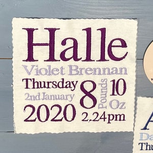 May include: Two white fabric squares with embroidered text. The top square says "Halle Violet Brennan Thursday 2nd January 2020 8 pounds 10 oz 2.24pm". The bottom square says "Alfie David Brennan Thursday 2nd January 2020 8 pounds 10 oz 2.24pm". A small round wooden sign with the astrological symbol for Scorpio and the text "www.memorgifts.com" is in the upper right corner.