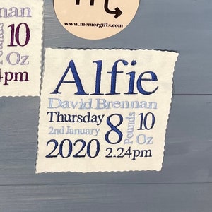 May include: Two white fabric squares with embroidered text. The top square says "Halle Violet Brennan Thursday 2nd January 2020 8 pounds 10 oz 2.24pm". The bottom square says "Alfie David Brennan Thursday 2nd January 2020 8 pounds 10 oz 2.24pm". A small round wooden sign with the astrological symbol for Scorpio and the text "www.memorgifts.com" is between the squares.