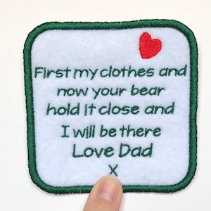 May include: A white felt square patch with green stitching around the edges. The patch has a red heart in the top right corner and the text "First my clothes and now your bear hold it close and I will be there Love Dad X" embroidered in black.