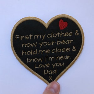 May include: Black heart-shaped patch with gold stitching and the text "First my clothes & now your bear hold me close & know I'm near. Love you Dad X" in gold thread. A small red heart is stitched in the upper right corner.