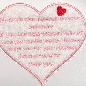 May include: Pink heart-shaped patch with a red heart at the top. The patch has pink embroidered text that reads: "My smile also depends on your behaviour. If you are aggressive I will not cure you I am like you I am human. Thank you for your respect. I am proud to help you."