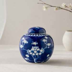 Op de afbeelding: Een kobaltblauwe keramische gemberpot met bijpassende deksel. De pot is versierd met witte bloempatronen. Een kleine witte vaas en een bloeiende tak staan op de achtergrond.