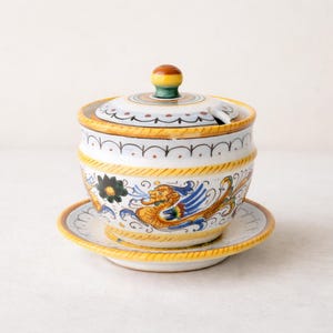 Op de afbeelding: Een keramische suikerpot met deksel en bijpassende schotel. De kom en schotel zijn wit met gele randen en handgeschilderde bloemen- en mythische wezensontwerpen in blauw, oranje en zwart. Het deksel heeft een kleine sleuf voor een lepel.