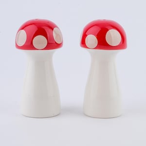 Op de afbeelding: Een paar paddenstoelvormige zout- en peperstrooiers. Elke strooier heeft een rode dop met witte stippen en een witte steel. De strooiers zijn van keramiek en hebben een glanzende afwerking. Ze zijn ontworpen om op een paddenstoel te lijken.