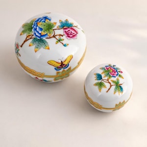 May include: Two white ceramic trinket boxes with floral and butterfly designs. The larger box features blue and pink flowers with a yellow butterfly. The smaller box has a similar floral design. Both have gold trim.