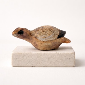 Può includere: Figurina di uccello in ceramica nei toni del marrone, beige e nero, posata su una base rettangolare bianco sporco. L'uccello ha un corpo dettagliato e strutturato e un piccolo occhio scuro. La figurina è un oggetto decorativo.