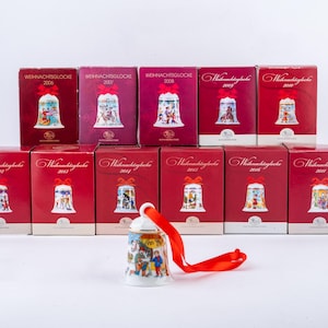 Könnte beinhalten: Eine Sammlung von Weihnachtsglocken-Ornamenten in roten Schachteln, jede mit einer dekorativen Glocke. Die Glocken haben farbenfrohe Illustrationen und sind mit dem Jahr beschriftet. Eine Glocke wird außerhalb ihrer Schachtel mit einem roten Band gezeigt.