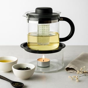 Puede incluir: Una tetera de vidrio con asa y tapa negras, llena de té y un infusor de té. La tetera se encuentra sobre un soporte de vidrio con una vela encendida debajo, con dos cuencos blancos pequeños y una cuchara cerca.