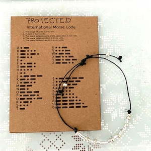 Peut inclure: Un bracelet en cordon noir avec des perles argentées. Le bracelet est noué et comporte une carte sur laquelle est imprimé l'alphabet et les chiffres du code Morse international. La carte indique "PROTECTED".