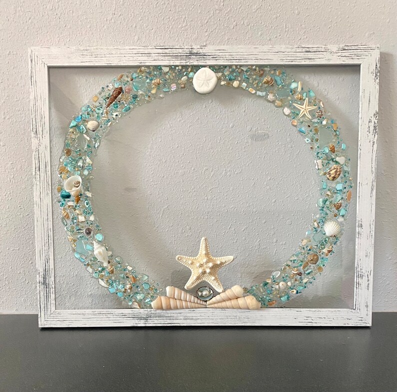 May include: A white framed wreath with a blue and white seashell design. The wreath is made of resin and includes seashells, sand, and a starfish.