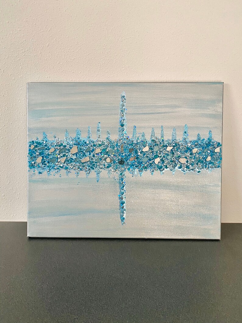 May include: Abstract artwork on canvas featuring a skyline design. The painting uses shades of blue and silver, with a textured, mosaic-like application of small, reflective pieces. The artwork is mounted on a rectangular canvas.