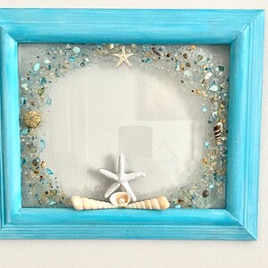 May include: A light blue picture frame with a white starfish and seashells embedded in the glass. The frame is decorated with a border of small blue and white glass pieces.
