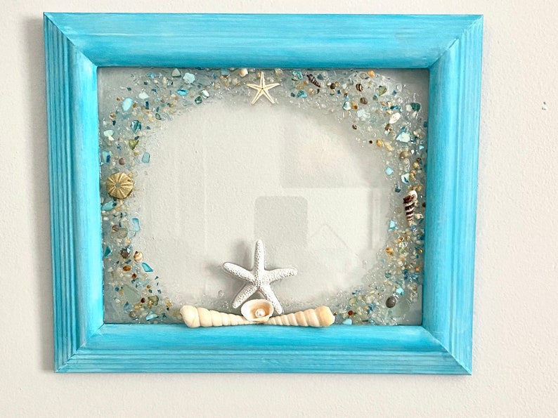 May include: A light blue picture frame with a white starfish and seashells embedded in the glass. The frame is decorated with a border of small blue and white glass pieces.