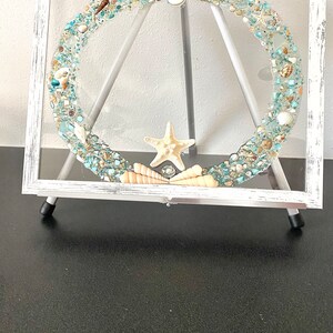 May include: A white framed picture with a clear glass insert filled with seashells, sea glass, and a starfish. The picture is on a white easel.