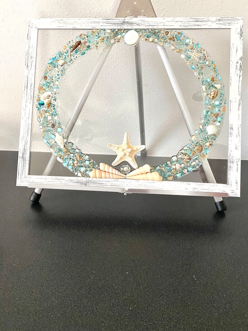 May include: A white framed picture with a clear glass insert filled with seashells, sea glass, and a starfish. The picture is on a white easel.
