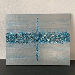 May include: Abstract canvas art with a silver and light blue background. The artwork features a central vertical line and a horizontal line, both adorned with blue and silver glass pieces, creating a textured, sparkling effect. The overall theme is modern and decorative.