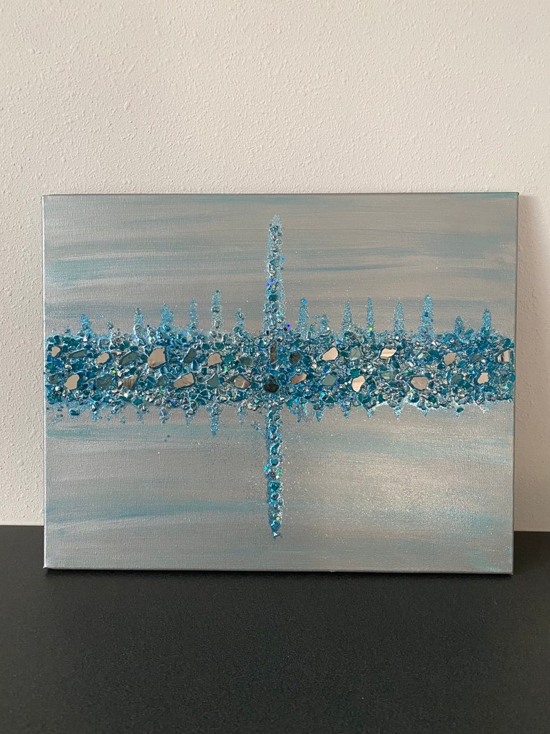 May include: Abstract canvas art with a silver and light blue background. The artwork features a central vertical line and a horizontal line, both adorned with blue and silver glass pieces, creating a textured, sparkling effect. The overall theme is modern and decorative.