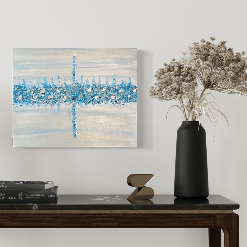 May include: Abstract canvas art featuring a horizontal band of blue and silver embellishments, with a vertical line intersecting it. The artwork is set against a light blue and cream background. A black vase with dried flowers and decorative items are on a dark table.