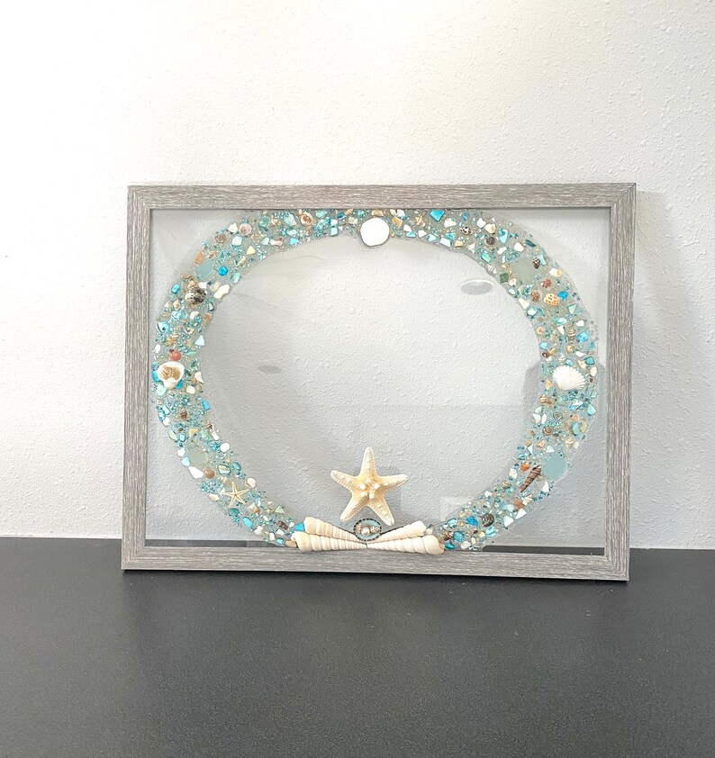 May include: A gray framed picture with a blue and white seashell wreath inside. The wreath is made of seashells, glass, and a starfish.