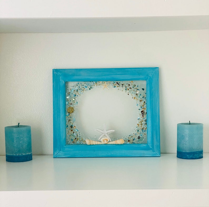 May include: A light blue picture frame with a white starfish and seashells inside. The frame is on a white shelf with two blue candles on either side.