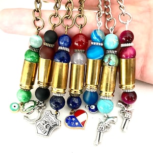May include: Seven keychains featuring bullet casings, each adorned with a different coloured bead and charm. The charms include a police badge, a heart with an American flag, an evil eye, and two guns.