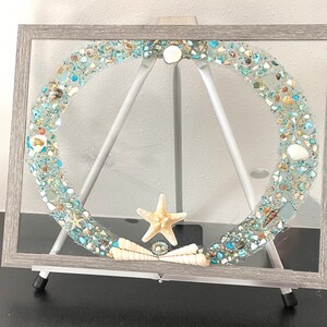 May include: A framed artwork featuring a circular design made of blue and white glass and seashells. The artwork is displayed on a wooden easel.
