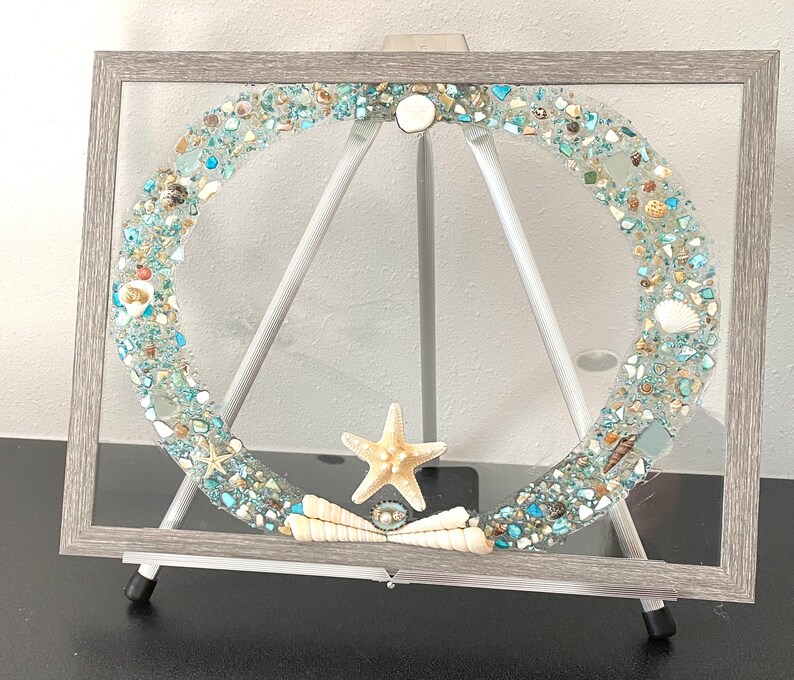 May include: A framed artwork featuring a circular design made of blue and white glass and seashells. The artwork is displayed on a wooden easel.