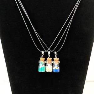 May include: Three glass bottle necklaces with cork stoppers and black cords. The bottles contain different colored sand, one blue, one white, and one green.