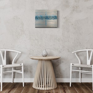 May include: Abstract square artwork with a silver background and a horizontal band of blue and silver accents. The artwork is mounted on a black frame and hangs on a gray wall. A small, round wooden table with a vase and two white chairs are in front of the wall.