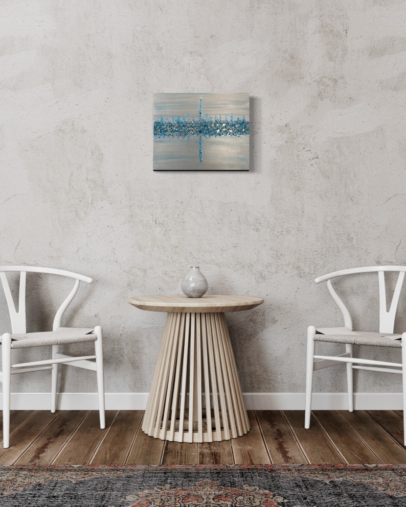 May include: Abstract square artwork with a silver background and a horizontal band of blue and silver accents. The artwork is mounted on a black frame and hangs on a gray wall. A small, round wooden table with a vase and two white chairs are in front of the wall.