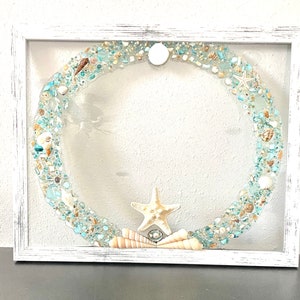 May include: A white framed square picture with a blue and white seashell wreath inside. The wreath is made of seashells, sea glass, and sand. A starfish is in the center of the wreath.