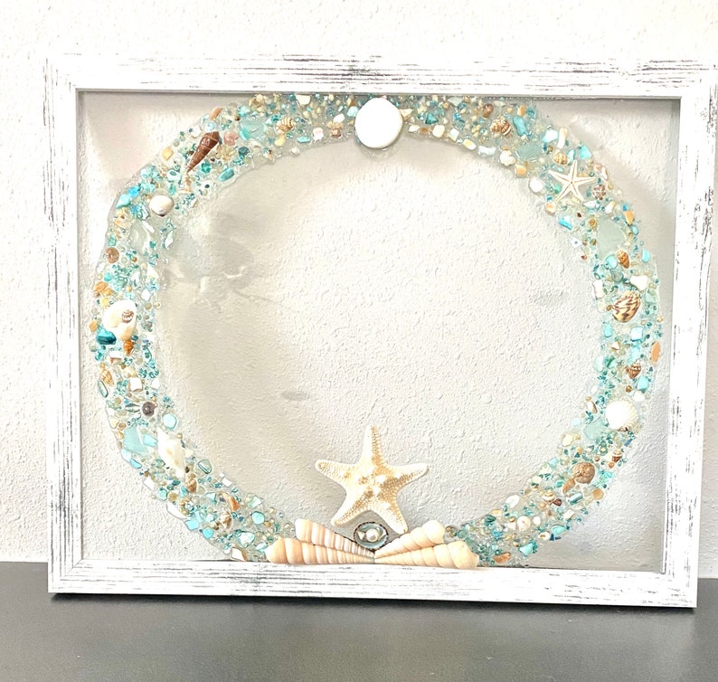 May include: A white framed square picture with a blue and white seashell wreath inside. The wreath is made of seashells, sea glass, and sand. A starfish is in the center of the wreath.