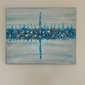 May include: Abstract canvas art with a light blue background and a central design of blue and iridescent stones. The design resembles a stylized cityscape or a cross, with vertical and horizontal elements.