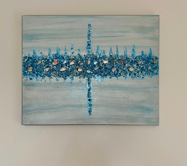 May include: Abstract canvas art with a light blue background and a central design of blue and iridescent stones. The design resembles a stylized cityscape or a cross, with vertical and horizontal elements.