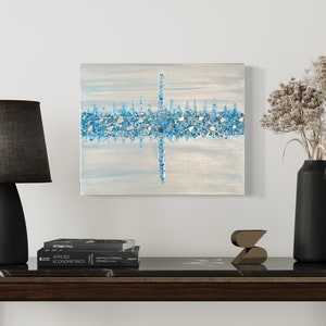 May include: An abstract painting with a light blue and white background, featuring a horizontal band of blue and silver mosaic-like elements. The artwork is displayed above a dark wooden table with a black lamp, books, and decorative items.