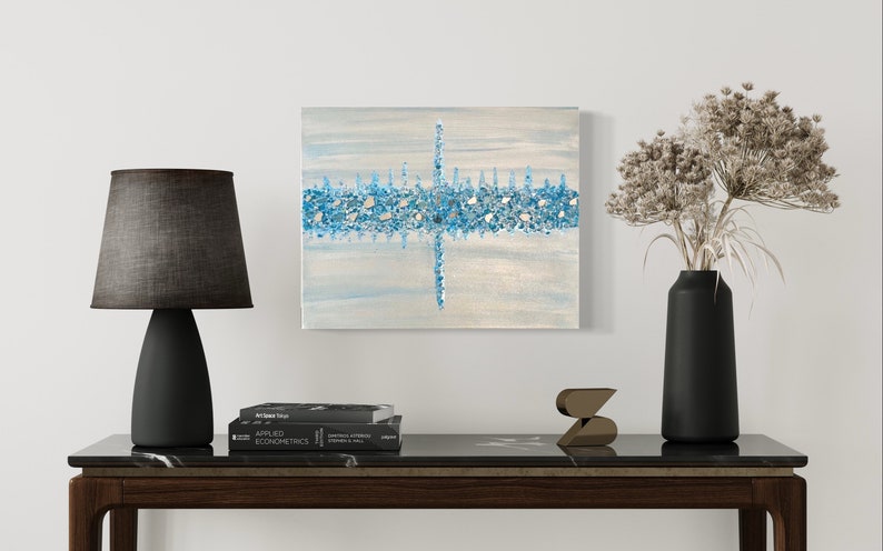 May include: An abstract painting with a light blue and white background, featuring a horizontal band of blue and silver mosaic-like elements. The artwork is displayed above a dark wooden table with a black lamp, books, and decorative items.