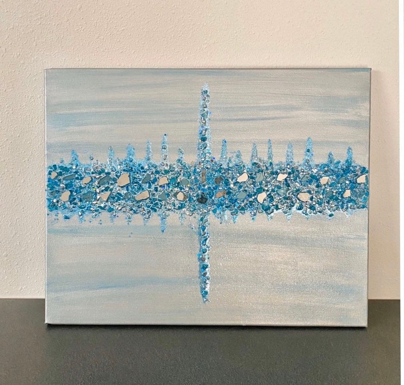 May include: Abstract canvas art featuring a light blue and silver background. A horizontal band of blue and silver glass pieces is centered on the canvas, with a vertical line of glass pieces intersecting it. The artwork has a textured appearance.