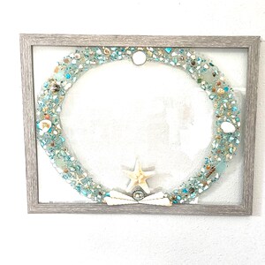 May include: A framed artwork featuring a circular design made of blue and white sea glass, shells, and a starfish. The frame is a light gray wood.
