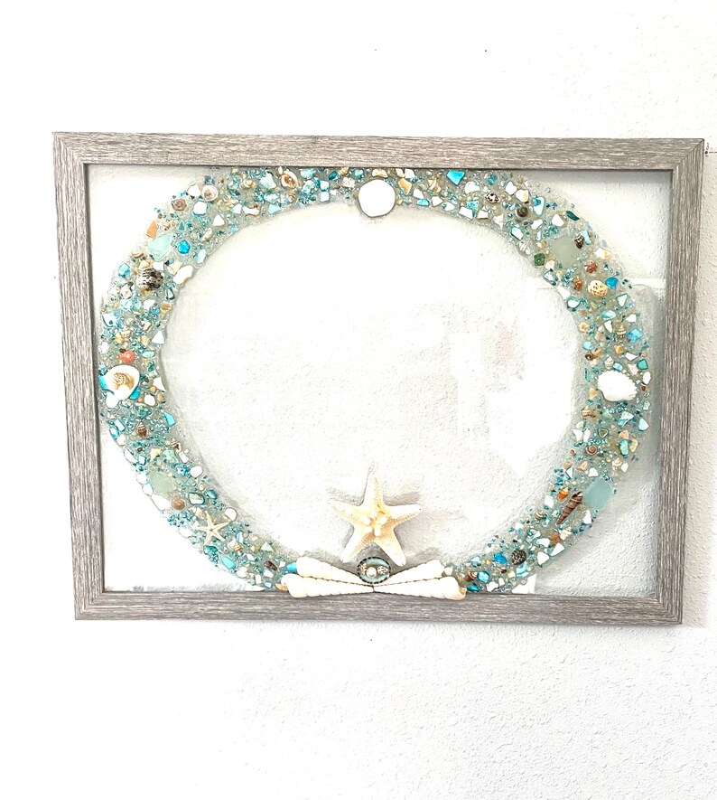 May include: A framed artwork featuring a circular design made of blue and white sea glass, shells, and a starfish. The frame is a light gray wood.