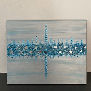 May include: A rectangular canvas art piece with a light blue and silver background. A horizontal band of blue and silver glass pieces is centered, with a vertical line of the same material intersecting it. The artwork is on a black surface.