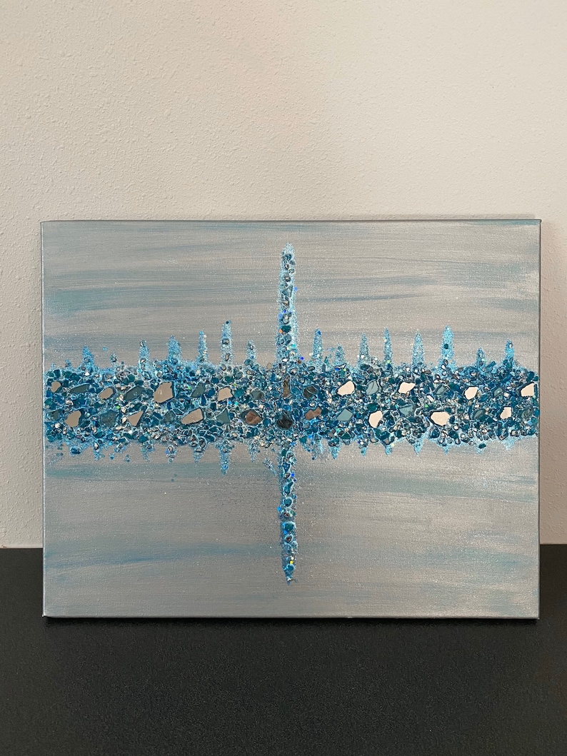 May include: A rectangular canvas art piece with a light blue and silver background. A horizontal band of blue and silver glass pieces is centered, with a vertical line of the same material intersecting it. The artwork is on a black surface.