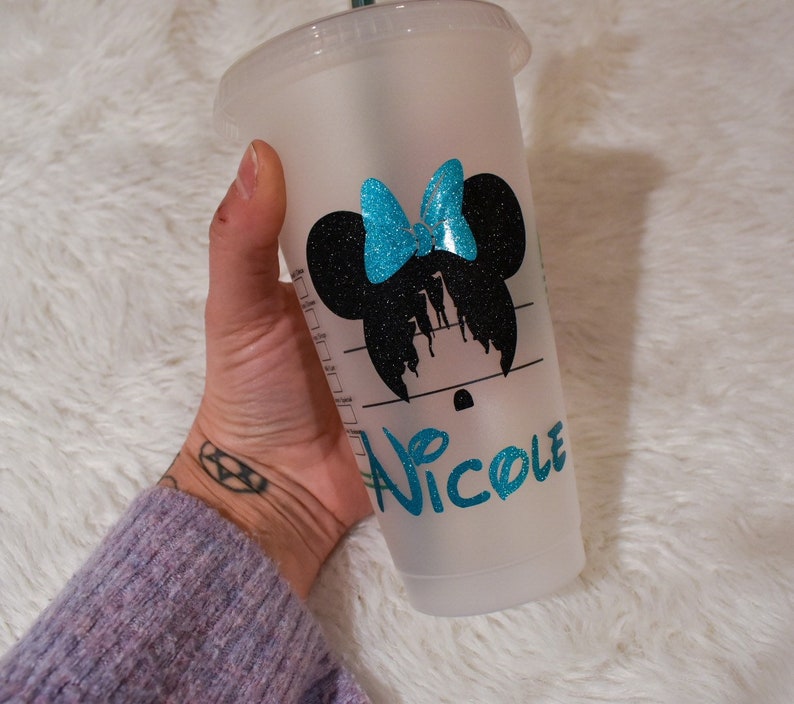 Puede incluir: Un vaso de pl&aacute;stico transparente de Starbucks con un dise&ntilde;o de silueta de Minnie Mouse negra y un lazo turquesa. El nombre "Nicole" est&aacute; escrito en gui&oacute;n turquesa. El vaso est&aacute; sostenido por una persona.
