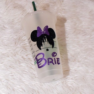 Puede incluir: Un vaso de pl&aacute;stico transparente para bebidas fr&iacute;as con tapa y pajita verde. El vaso presenta una silueta negra de Minnie Mouse con un lazo morado y el nombre "Brie" en letras con purpurina morada. Sobre una superficie blanca y esponjosa.