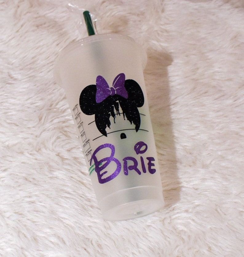 Puede incluir: Un vaso de pl&aacute;stico transparente para bebidas fr&iacute;as con tapa y pajita verde. El vaso presenta una silueta negra de Minnie Mouse con un lazo morado y el nombre "Brie" en letras con purpurina morada. Sobre una superficie blanca y esponjosa.