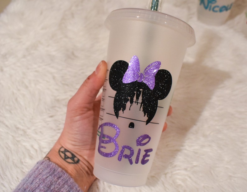 Puede incluir: Un vaso fr&iacute;o de pl&aacute;stico transparente con un dise&ntilde;o de Minnie Mouse con purpurina negra y un lazo de purpurina morada. El nombre "Brie" est&aacute; escrito en purpurina morada en el vaso. El vaso tiene una pajita verde.