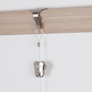 May include: Silver metal hook for hanging a curtain rod, with a silver metal clip attached to a clear cord hanging from the hook.