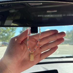 May include: A hanging car accessory featuring a string of small, multicolored translucent beads arranged in a repeating pattern. A single, pale yellow, semi-translucent gummy bear charm is attached to the center of the beaded loop.