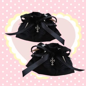 May include: Two black drawstring bags with silver spikes, lace trim, and a silver cross charm. Each bag has a large black bow. The bags are set against a pink background with white polka dots and a yellow and white scalloped border.